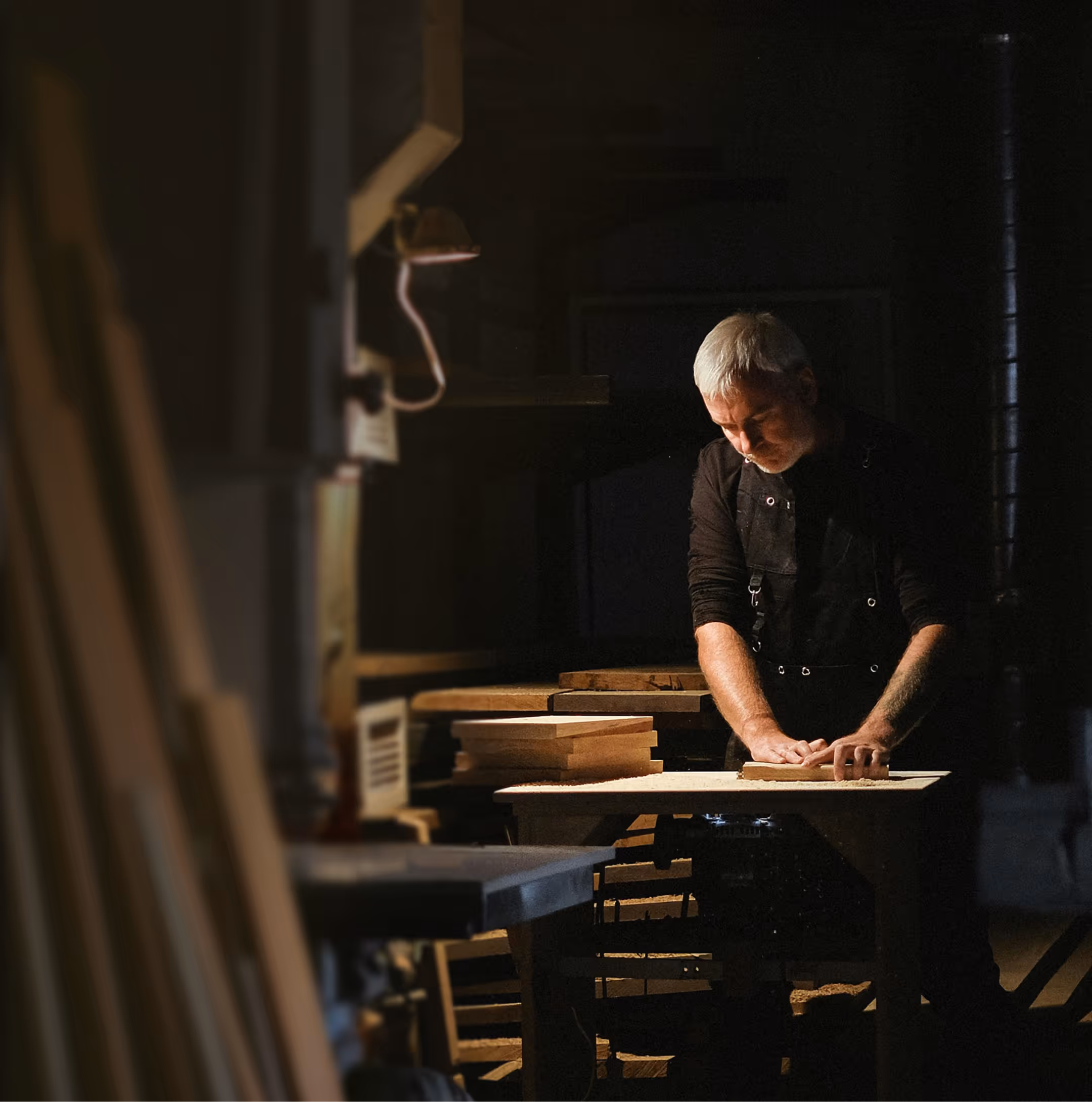 Man with gray hair in a dimly lit workshop, sanding or working on a piece of wood on a table.